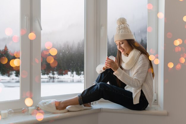 Mujer atractiva joven en elegante suéter de punto blanco, bufanda y sombrero sentado en casa en el alféizar de la ventana en Navidad con decoración de presente de bola de nieve de cristal, vista del bosque de invierno, luces bokeh