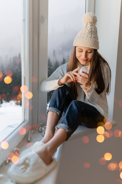 Mujer atractiva joven en elegante suéter de punto blanco, bufanda y sombrero sentado en casa en el alféizar de la ventana en Navidad con decoración de presente de bola de nieve de cristal, vista del bosque de invierno, luces bokeh