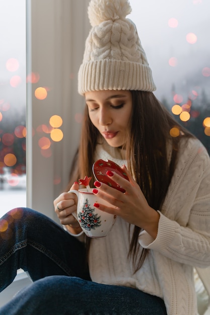 Mujer atractiva joven en elegante suéter de punto blanco, bufanda y sombrero sentado en casa en el alféizar de la ventana en Navidad sosteniendo una taza bebiendo té caliente, vista de fondo de bosque de invierno, luces bokeh
