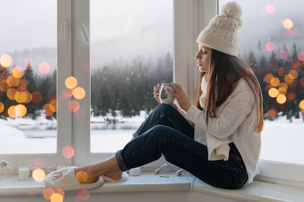 Mujer atractiva joven en elegante suéter de punto blanco, bufanda y sombrero sentado en casa en el alféizar de la ventana en Navidad sosteniendo una taza bebiendo té caliente, vista de fondo de bosque de invierno, luces bokeh