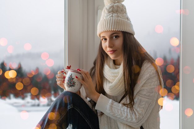 Mujer atractiva joven en elegante suéter de punto blanco, bufanda y sombrero sentado en casa en el alféizar de la ventana en Navidad sosteniendo una taza bebiendo té caliente, vista de fondo de bosque de invierno, luces bokeh