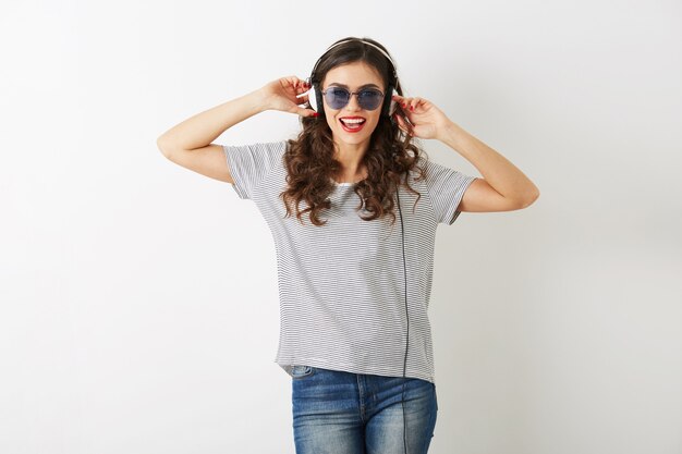 Mujer atractiva joven escuchando música en auriculares, con gafas de sol, cabello rizado, humor lúdico, aislado sobre fondo blanco, camiseta, estilo hipster casual, feliz emoción positiva, emocional