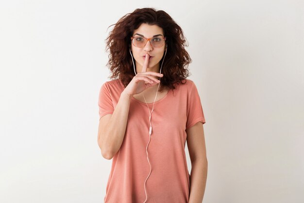 Mujer atractiva joven en gafas escuchando música en los auriculares, sosteniendo el dedo en los labios, mostrando gesto de silencio, divertida emoción sorprendida, cabello rizado, camiseta aislada, rosa