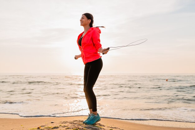 Mujer atractiva joven haciendo ejercicios deportivos en el amanecer de la mañana en la playa del mar en ropa deportiva, estilo de vida saludable, escuchando música con auriculares, vistiendo una chaqueta cortavientos rosa, saltando en la cuerda