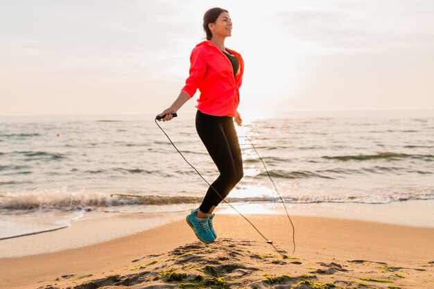 Mujer atractiva joven haciendo ejercicios deportivos en el amanecer de la mañana en la playa del mar en ropa deportiva, estilo de vida saludable, escuchando música con auriculares, vistiendo una chaqueta cortavientos rosa, saltando en la cuerda