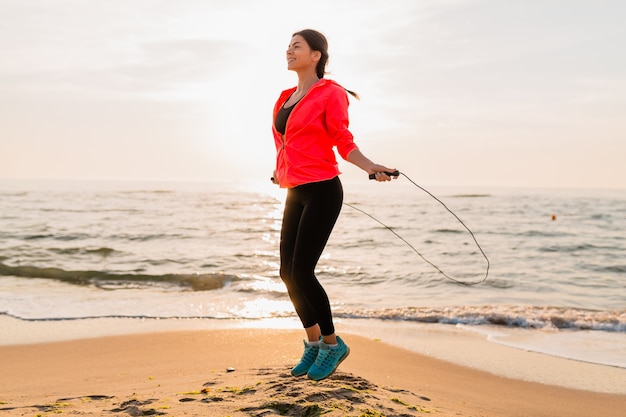 Mujer atractiva joven haciendo ejercicios deportivos en el amanecer de la mañana en la playa del mar en ropa deportiva, estilo de vida saludable, escuchando música con auriculares, vistiendo una chaqueta cortavientos rosa, saltando en la cuerda