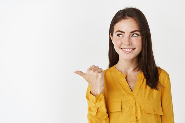 Mujer atractiva joven mirando la oferta promocional, apuntando con el pulgar hacia la izquierda y mirando la pancarta con una sonrisa satisfecha, de pie en la pared blanca