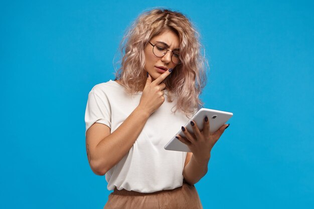 Mujer atractiva joven pensativa navegando por Internet leyendo noticias del mundo o revisando el correo electrónico en la tableta digital. Linda chica pensativa en gafas usando una computadora portátil con panel táctil genérico para trabajo remoto