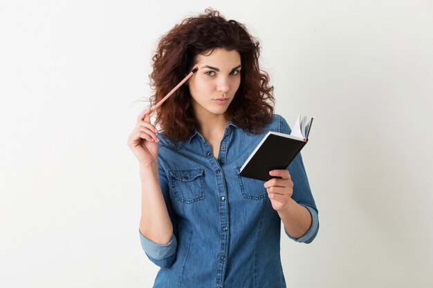 Mujer atractiva joven que sostiene el cuaderno y el lápiz, pensando, expresión seria de la cara, pelo rizado, pensativo, aislado, camisa azul del dril de algodón, aprendizaje del estudiante, educación