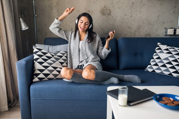 Mujer atractiva joven en ropa casual relajante en casa, tiempo libre, divertirse, sonriendo escuchando música en auriculares, bailando sentado en el sofá, alegre, feliz, con medias, suéter