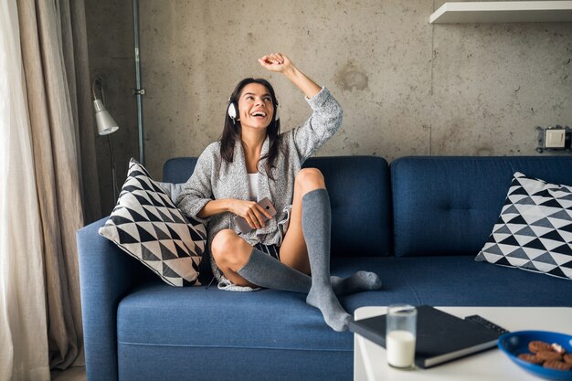 Mujer atractiva joven en ropa casual relajante en casa, tiempo libre, divertirse, sonriendo escuchando música en auriculares, bailando sentado en el sofá, alegre, feliz, con medias, suéter