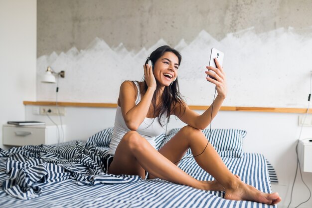 Mujer atractiva joven sentada en la cama en pijama, sonriendo en el dormitorio, emoción feliz, despertarse por la mañana, riendo, sexy, flaca, escuchando música en auriculares, usando el teléfono inteligente, tomando fotos selfie