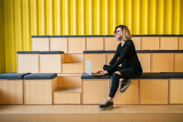 Mujer atractiva joven sentada en la sala de conferencias, trabajando en la computadora portátil, con gafas, auditorio moderno, educación de estudiantes en línea, autónomo, sonriente, inicio adolescente, mirando a la cámara, feliz