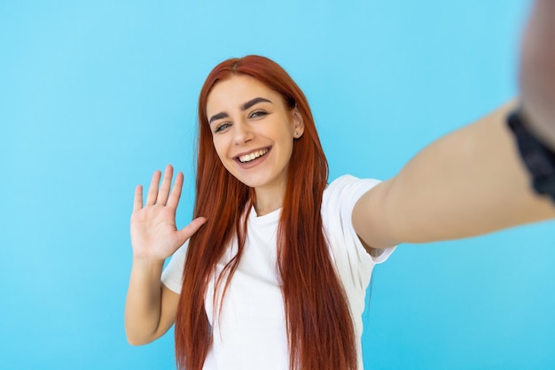 Mujer atractiva joven tiene saludo de onda de videollamada en la cámara de su teléfono aislado en azul