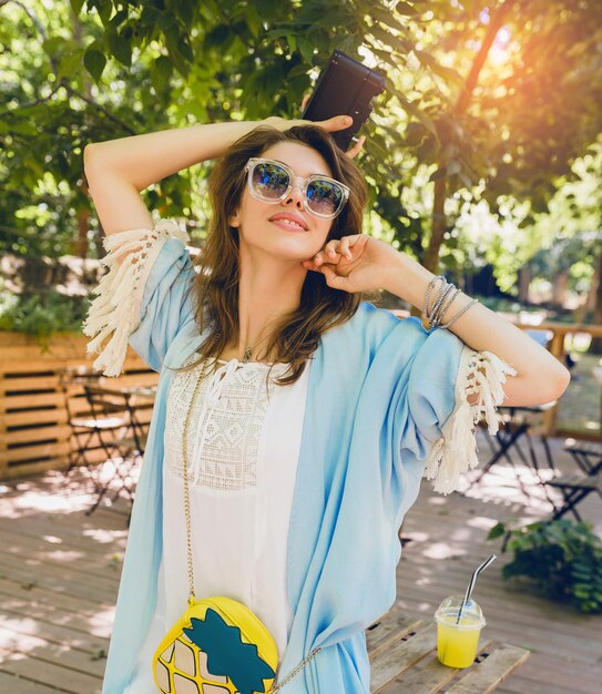 Mujer atractiva joven en traje de moda de verano, estilo hipster, vestido blanco, capa azul, bolso amarillo, gafas de sol, sonriendo, sosteniendo una cámara de fotos vintage, accesorios elegantes, ropa de moda