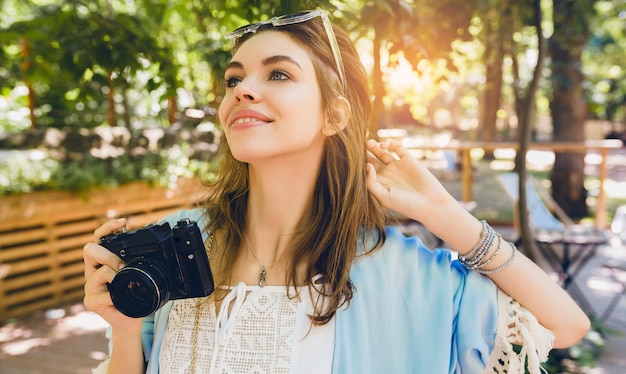 Mujer atractiva joven en traje de moda de verano, estilo hipster, vestido blanco, capa azul, gafas de sol, sonriendo, sosteniendo una cámara de fotos vintage, accesorios elegantes, relajándose en vacaciones, ropa de moda