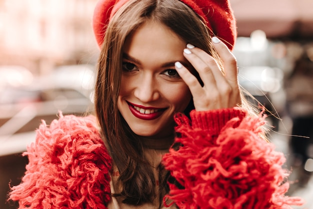 Mujer atractiva con labios brillantes sonriendo y tocando su cabello oscuro. Closeup retrato de dama en traje rojo y tocado.