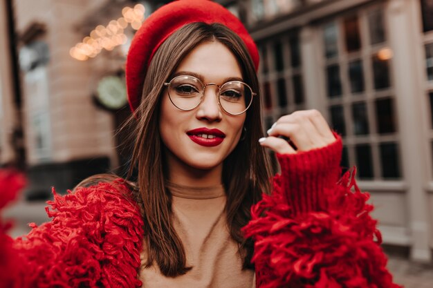 Mujer atractiva con labios rojos hace selfie en la calle. Foto de morena con gafas vestidas con elegante sombrero, chaqueta roja y top beige.