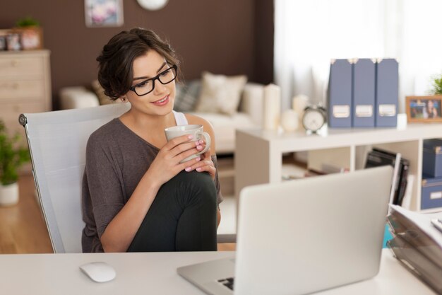 Mujer atractiva mirando en la computadora portátil mientras toma café en casa