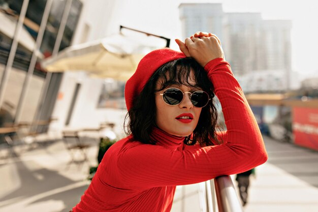 Mujer atractiva de moda con cabello corto y labios rojos con gorra roja y camisa roja está tocando la cabeza y sonriendo a la cámara mientras camina por la ciudad