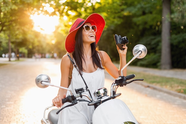 Mujer atractiva montando en moto en la calle, estilo de vacaciones de verano, viajando, sonriendo, feliz, divirtiéndose, atuendo elegante, aventuras, tomando fotos en la cámara de fotos vintage