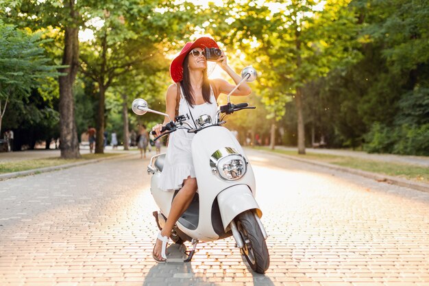 Mujer atractiva montando en moto en la calle, estilo de vacaciones de verano, viajando, sonriendo, feliz, divirtiéndose, atuendo elegante, aventuras, tomando fotos en la cámara de fotos vintage