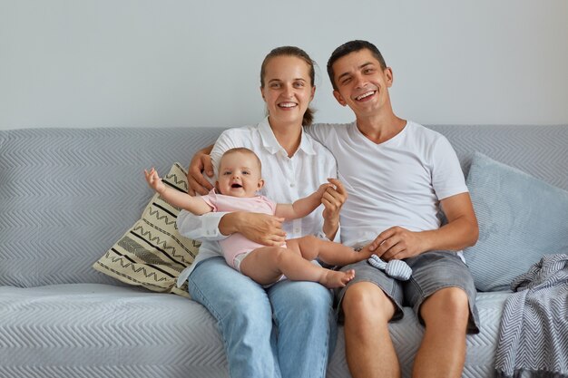 Mujer atractiva y mujer hermosa sentada en el sofá con la hija pequeña, mirando sonriendo a la cámara, siendo felices juntos, familia en casa, tiro interior.