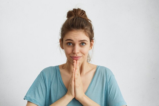 Mujer atractiva con ojos azules y piel sana con expresión de esperanza manteniendo sus manos juntas pidiendo algo de esperanza.