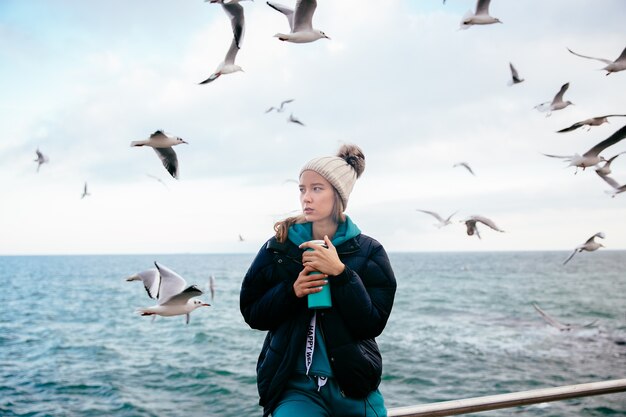 Mujer atractiva pensando en algo, sostiene termo cerca del océano con gaviotas