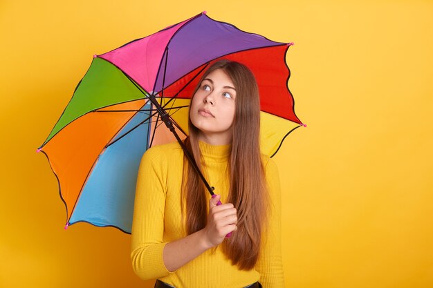 Mujer atractiva pensativa sosteniendo paraguas multicolor y mirando a un lado, chica con cabello largo y hermoso