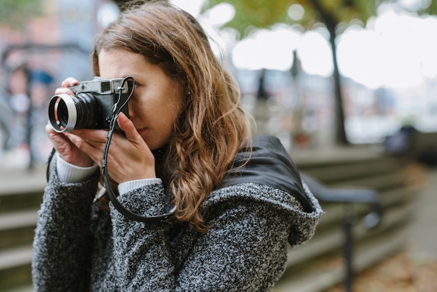 Mujer atractiva de pie vistiendo un abrigo gris y tomando una foto con una cámara vintage