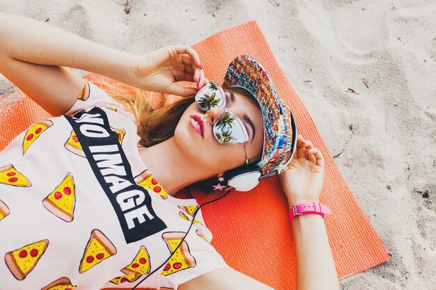 Mujer atractiva en la playa escuchando música en auriculares con elegante traje colorido en vacaciones tropicales de verano con gafas de sol de gorra de accesorios, sonriendo feliz acostado en la estera de yoga vista desde arriba