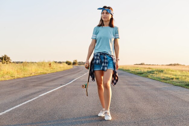 Mujer atractiva positiva vistiendo pantalones cortos, camiseta y banda para el cabello sosteniendo el patín en las manos y mirando a otro lado, caminando por la carretera asfaltada en la puesta de sol en verano.