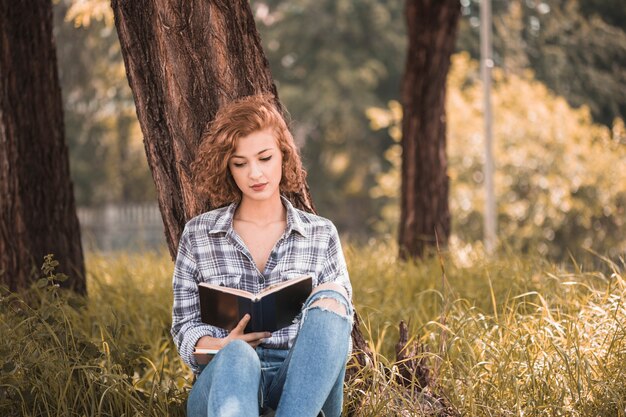 Mujer atractiva que se inclina en árbol y libro de lectura en jardín público