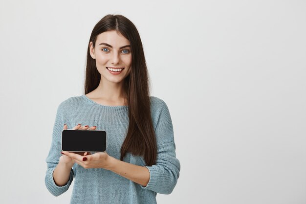 Mujer atractiva que muestra la pantalla del teléfono inteligente y sonriendo, recomienda la aplicación