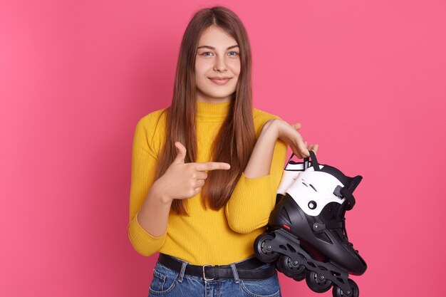 Mujer atractiva que sostiene el patinaje en manos y que lo señala con su dedo índice, posando contra la pared rosada.