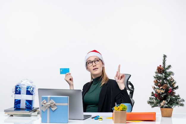 Mujer atractiva segura con sombrero de santa claus y gafas sentado en una mesa y sosteniendo una tarjeta bancaria y apuntando hacia arriba en la oficina