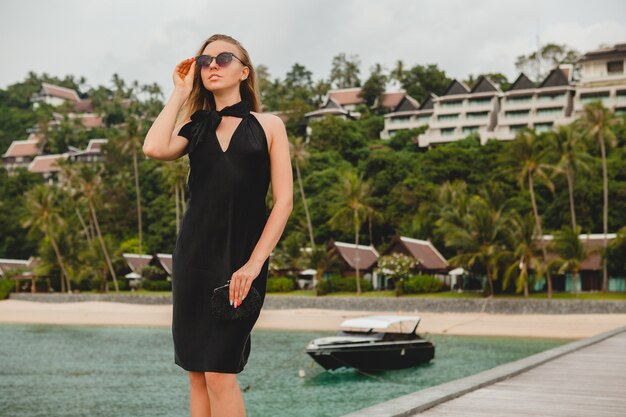 Mujer atractiva sexy de lujo vestida con vestido negro posando en el muelle en el hotel resort de lujo, con gafas de sol, vacaciones de verano, playa tropical