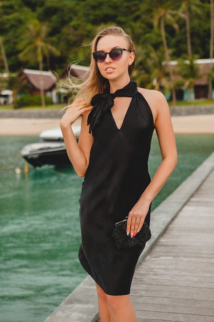 Mujer atractiva sexy de lujo vestida con vestido negro posando en el muelle en el hotel resort de lujo, con gafas de sol, vacaciones de verano, playa tropical