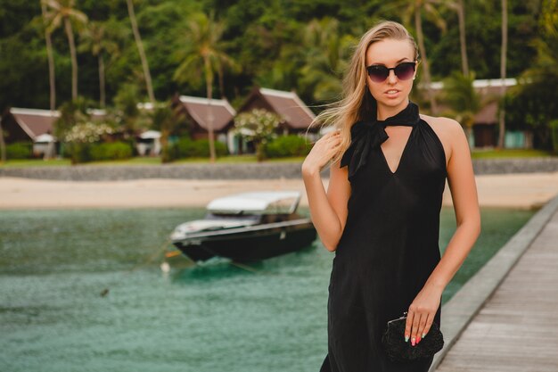 Mujer atractiva sexy de lujo vestida con vestido negro posando en el muelle en el hotel resort de lujo, con gafas de sol, vacaciones de verano, playa tropical