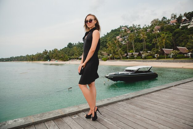 Mujer atractiva sexy de lujo vestida con vestido negro posando en el muelle en el hotel resort de lujo, con gafas de sol, vacaciones de verano, playa tropical