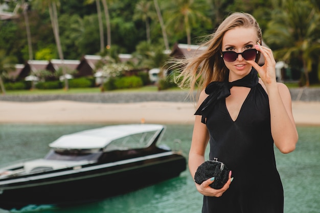 Mujer atractiva sexy de lujo vestida con vestido negro posando en el muelle en el hotel resort de lujo, con gafas de sol, vacaciones de verano, playa tropical