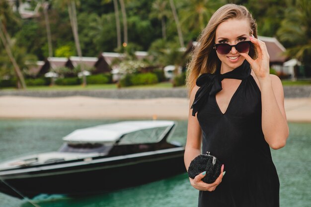 Mujer atractiva sexy de lujo vestida con vestido negro posando en el muelle en el hotel resort de lujo, con gafas de sol, vacaciones de verano, playa tropical