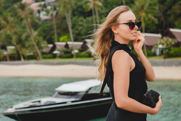 Foto gratuita mujer atractiva sexy de lujo vestida con vestido negro posando en el muelle en el hotel resort de lujo, con gafas de sol, vacaciones de verano, playa tropical