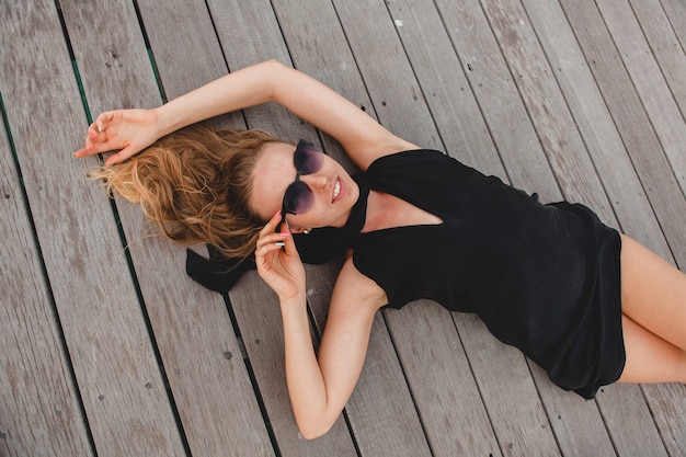 Mujer atractiva sexy de lujo vestida con vestido negro tirado en el suelo con gafas de sol, vacaciones de verano, vista desde arriba
