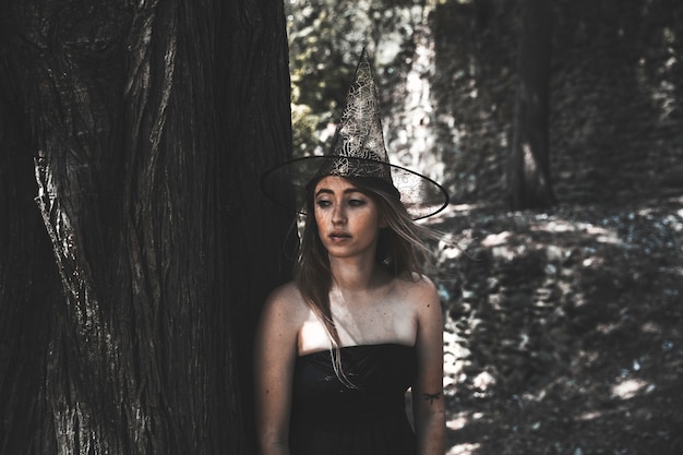 Mujer atractiva en el sombrero de bruja al lado del árbol y mirando lejos