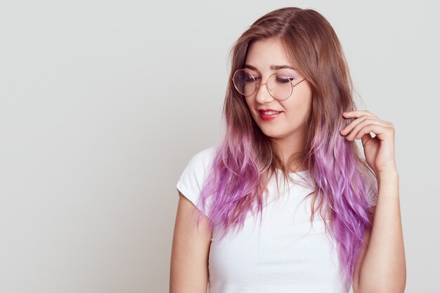 Mujer atractiva soñadora con cabello lila mirando a otro lado y tocándose el cabello, vestida con camiseta blanca y gafas, copia espacio para publicidad, posando aislada sobre pared gris.