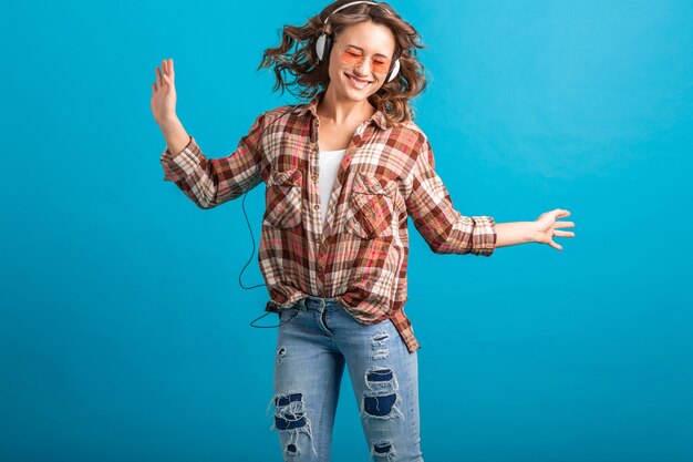 Mujer atractiva sonriendo disfrutando de escuchar música en auriculares con camisa a cuadros y jeans aislado sobre fondo azul de estudio, con gafas de sol rosa