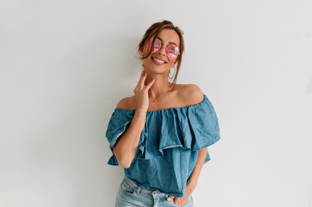 Mujer atractiva sonriente con gafas de sol tocándose la barbilla y mirando a un lado sobre un fondo aislado Retrato interior de una mujer bronceada europea