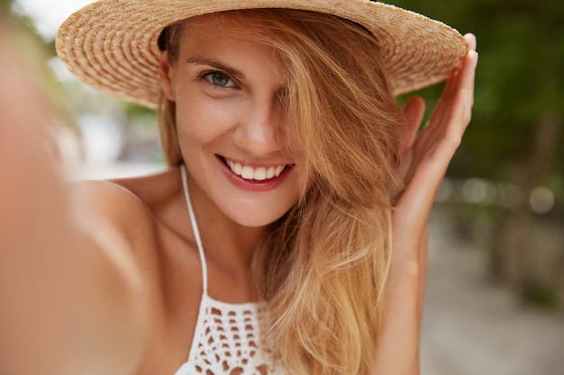 Mujer atractiva con sonrisa agradable, cabello claro, usa sombrero de verano, hace selfie con dispositivo irreconocible mientras camina al aire libre, disfruta de hermosos paisajes y clima cálido y brillante. Mujer alegre